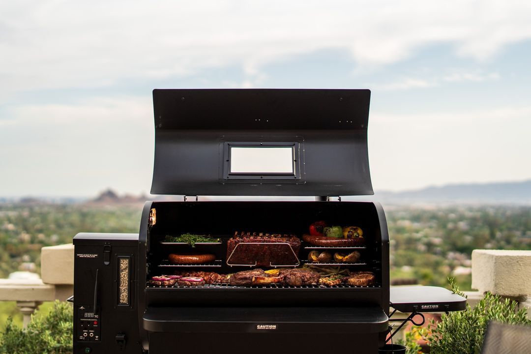 A grill with the lid open is cooking food on a patio.