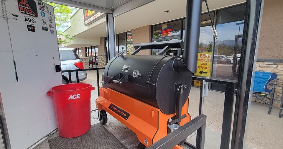 A large grill is sitting outside of a building next to a red trash can.