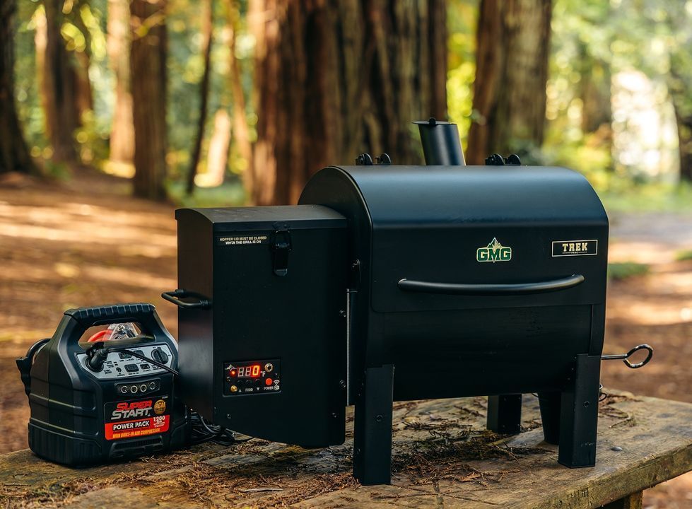 A wood pellet grill is sitting on top of a wooden table in the woods.
