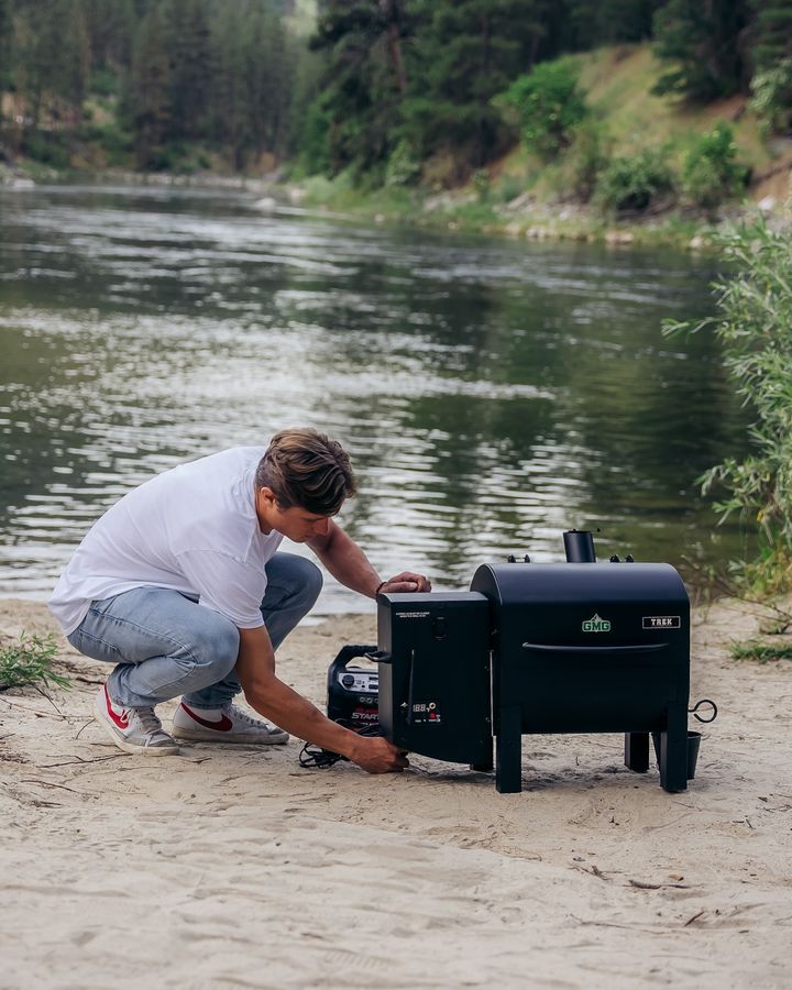 A man is kneeling down next to a grill on the beach near a river.