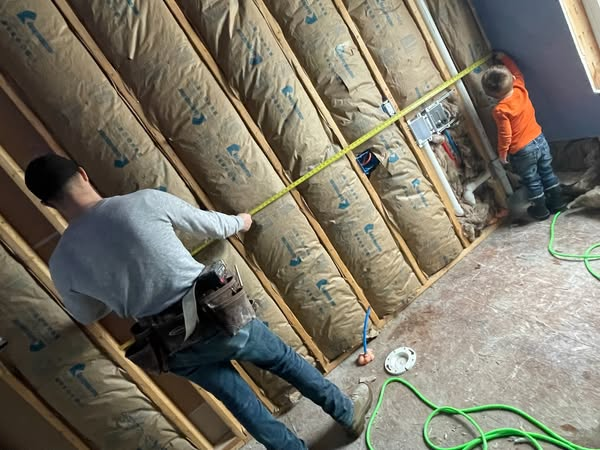 Two people measuring insulation in a wall; one holds the tape measure. Interior setting with wooden beams.