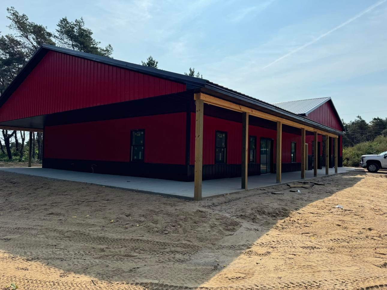 Red and black metal building with a porch, on a sandy lot, with a pickup truck in the background.