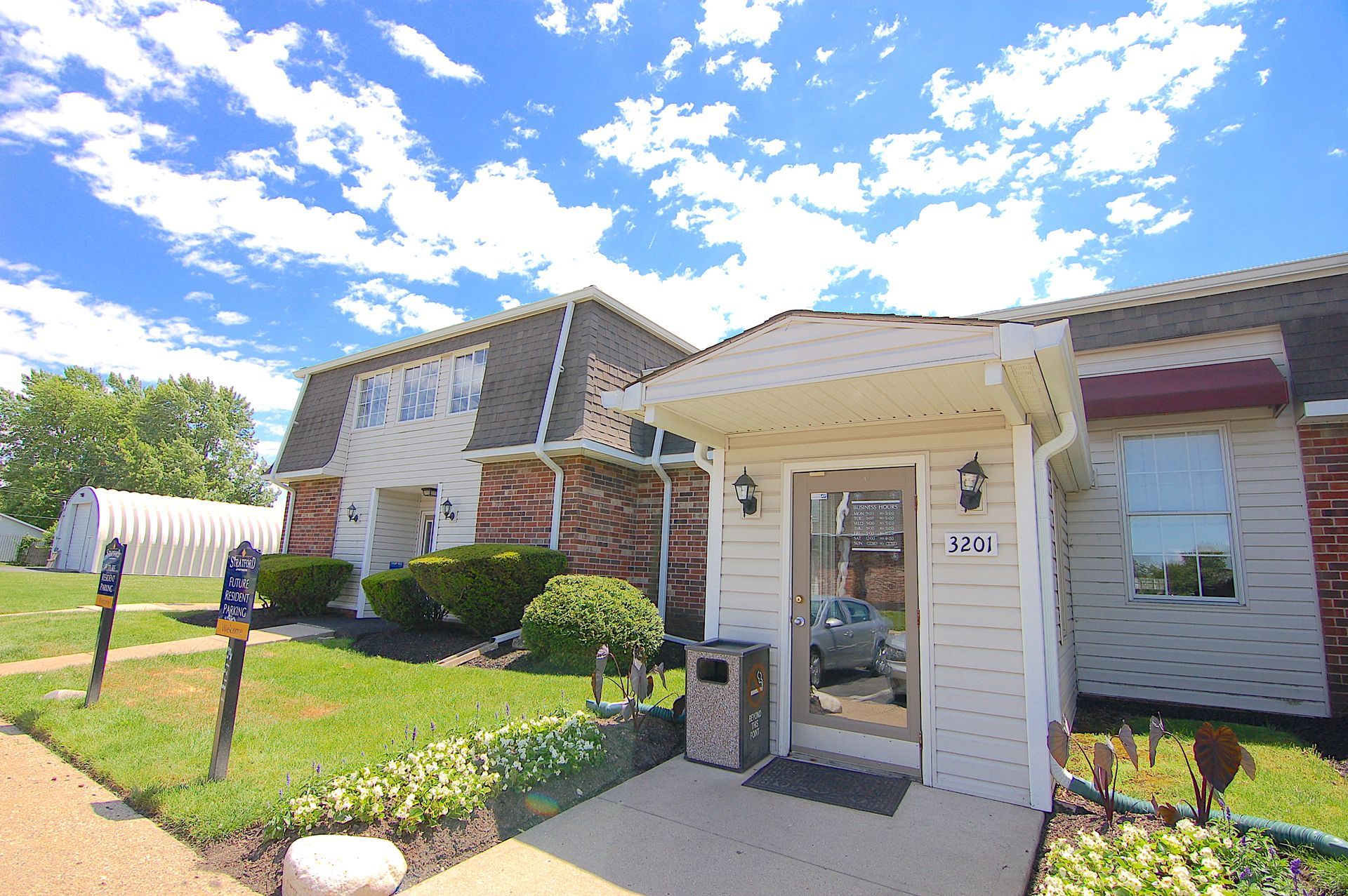 Residential building entrance with white siding, glass door, and landscaped lawn under a bright blue sky.