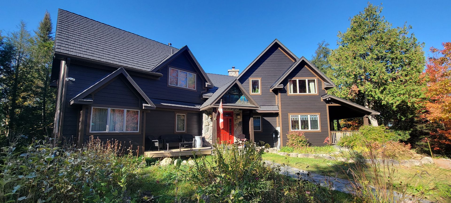 Dark wooden house with steep roofs and red door, surrounded by trees and wildflowers under a blue sky