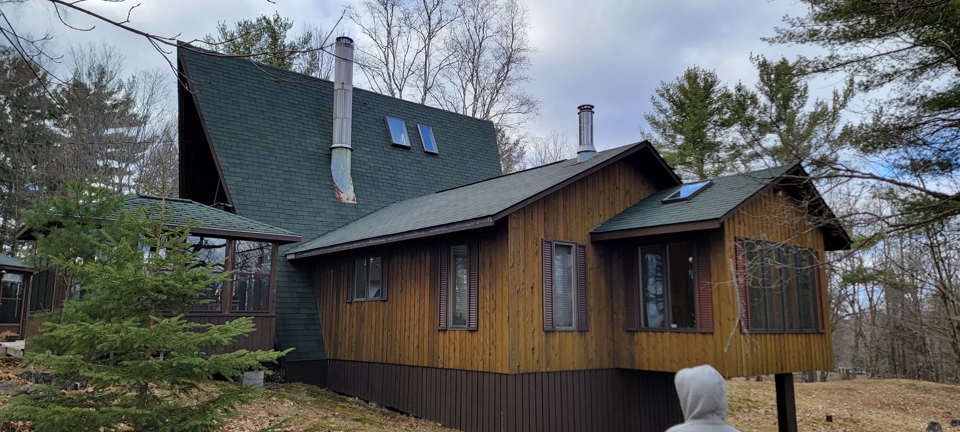 A person is standing in front of a wooden house with a green roof.