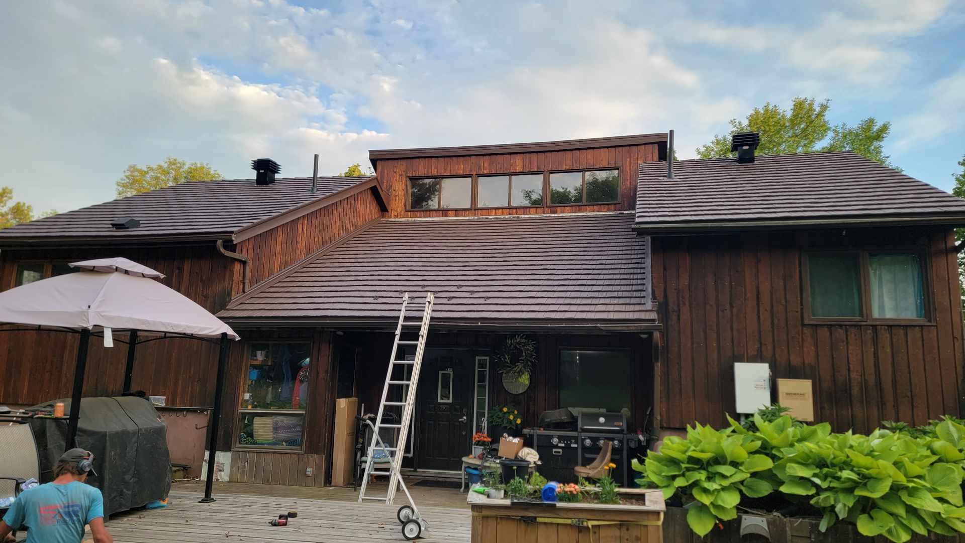 A man is sitting under an umbrella in front of a wooden house.
