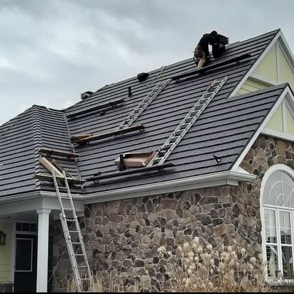 A person working on a dark gray shingled roof of a house with stone siding, ladders present.