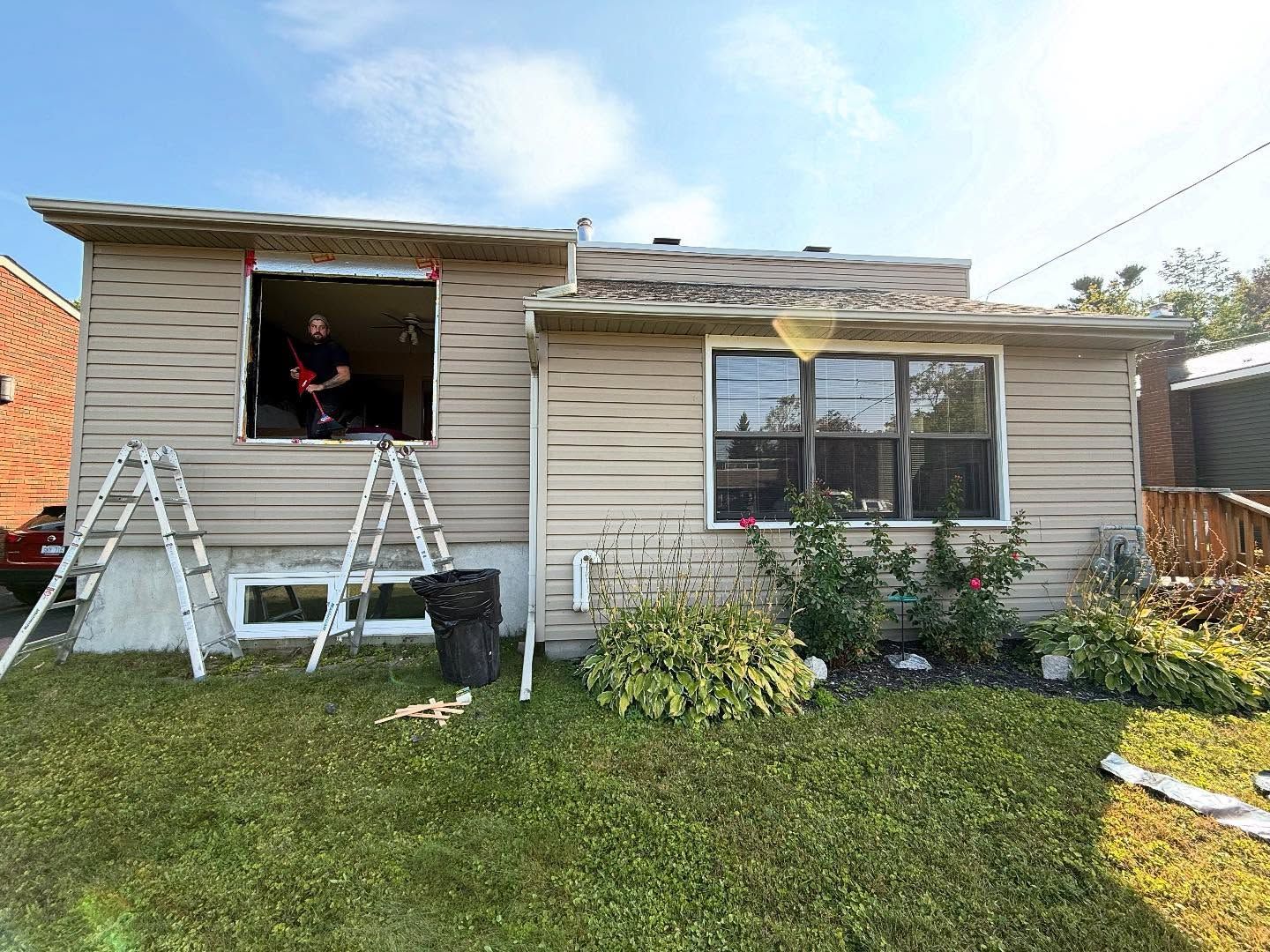 A person works on a house exterior, removing a window. Two ladders stand near the opening.