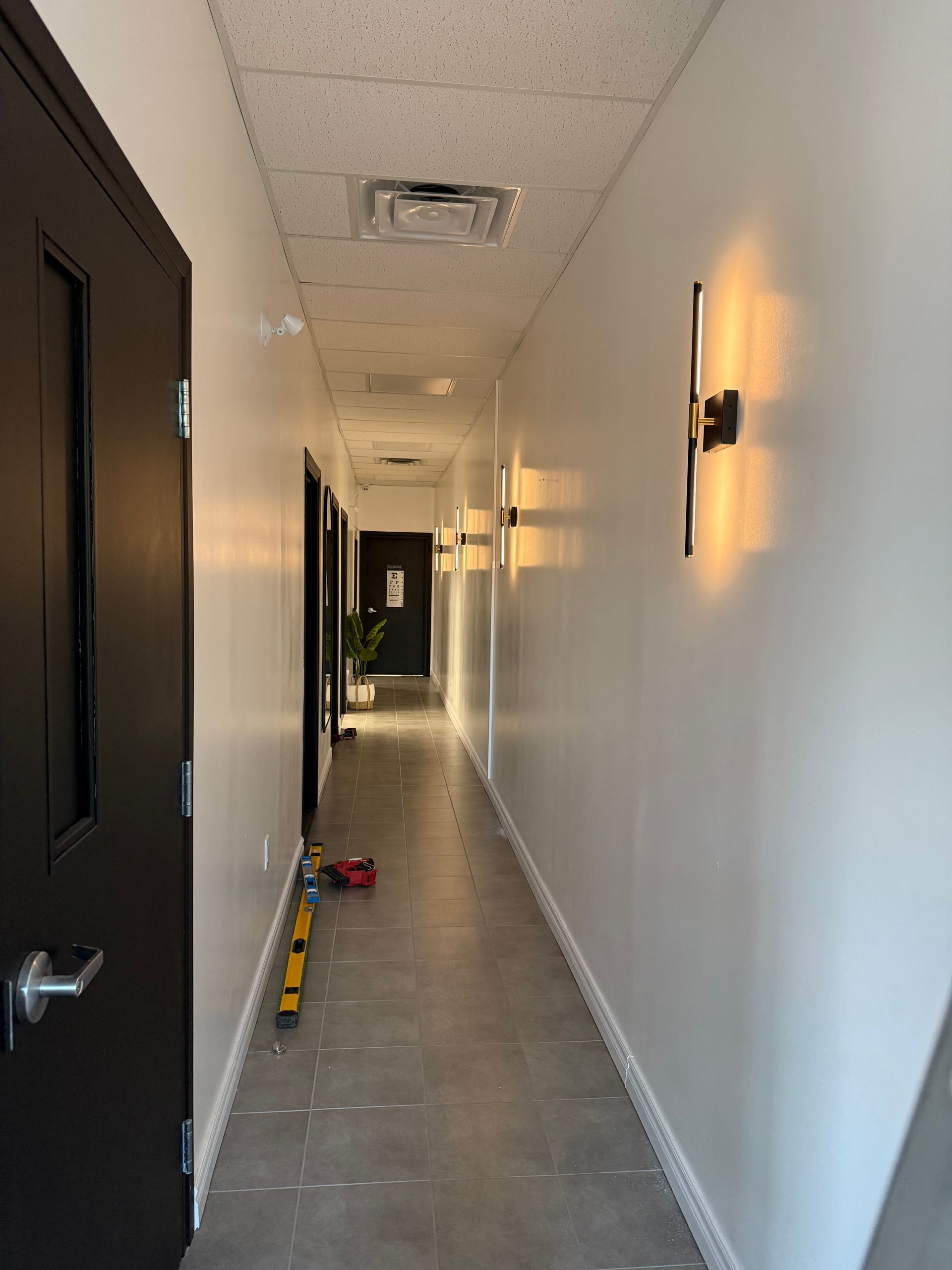 Long apartment hallway with white walls, black doors, wall lights, and a vacuum and tools on the floor.