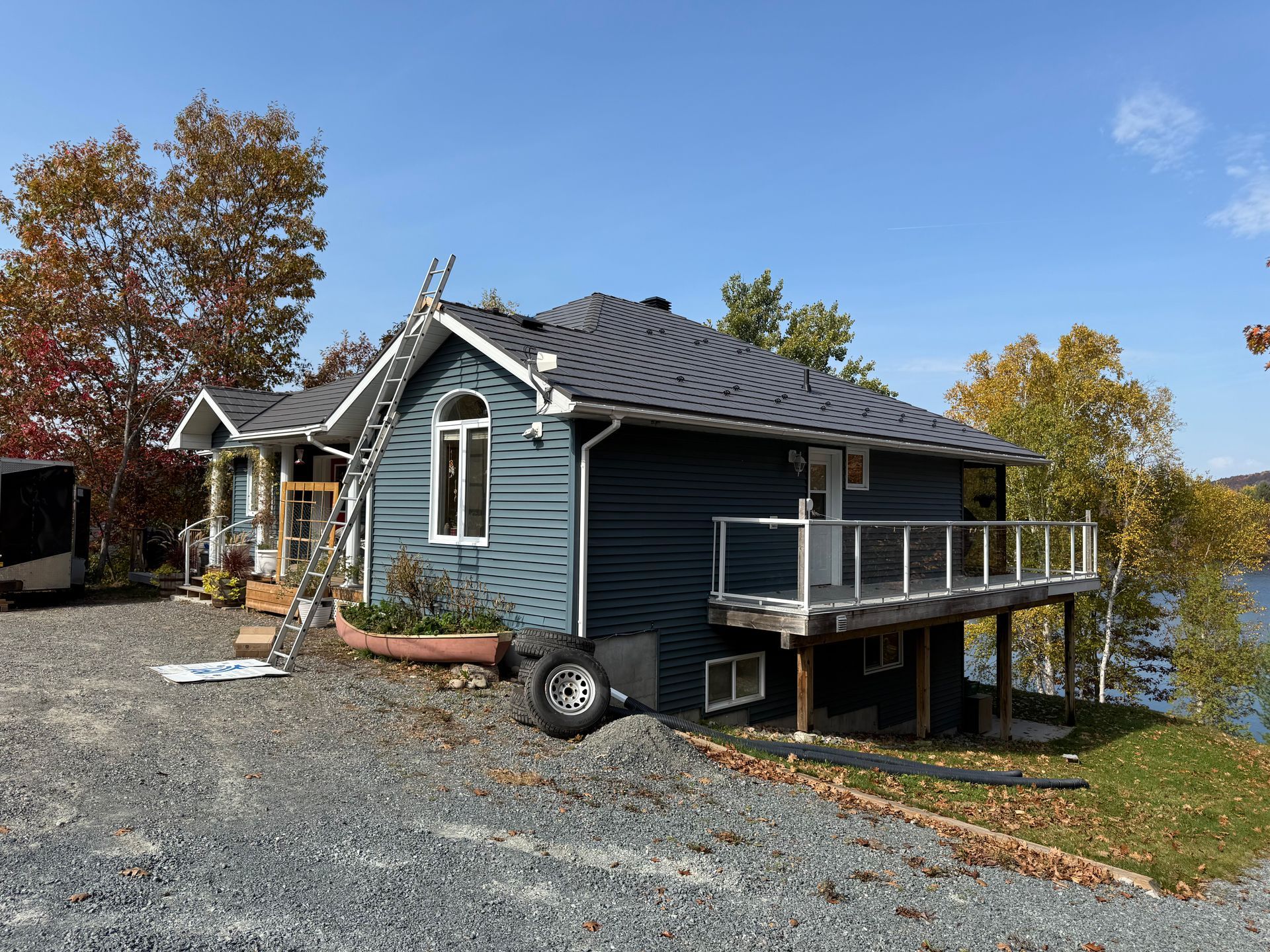 A blue house with a dark roof and deck sits on a gravel lot near trees and water, with a ladder leaning against the side.
