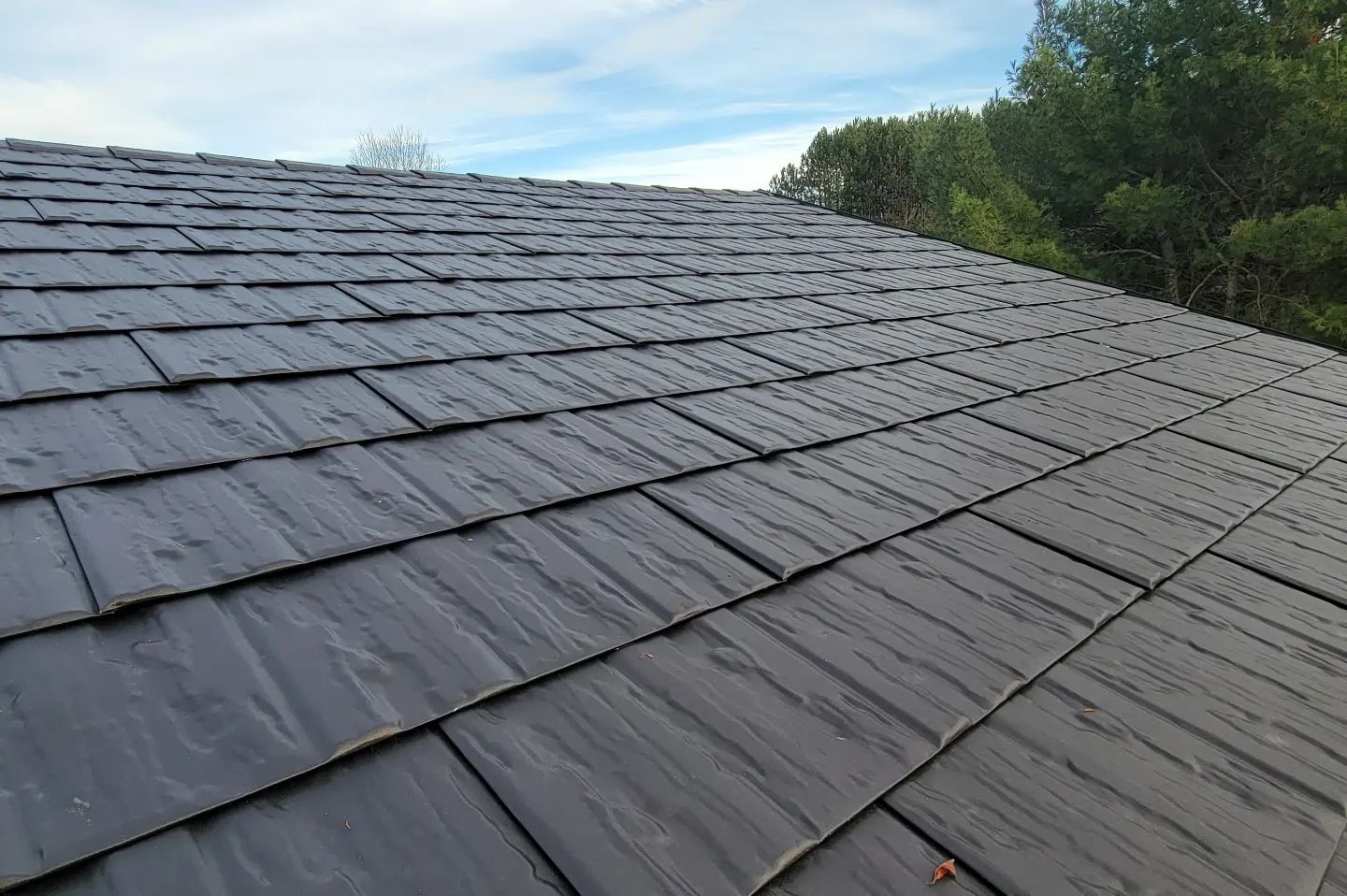 A close-up of a dark grey, textured shingled roof under a clear blue sky with trees visible in the distance.