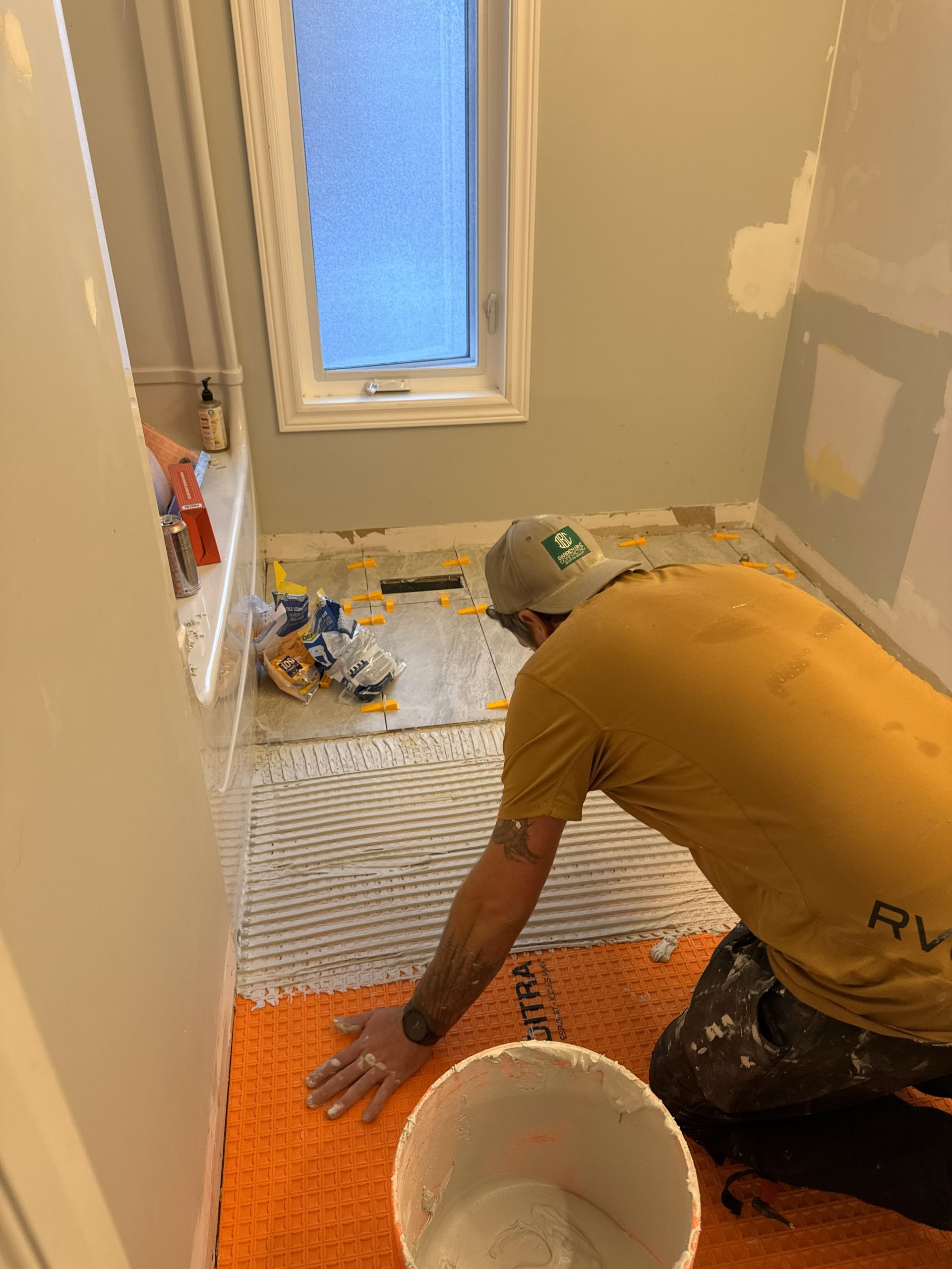 Person tiling a bathroom floor, orange mat, gray walls, window, white bucket.