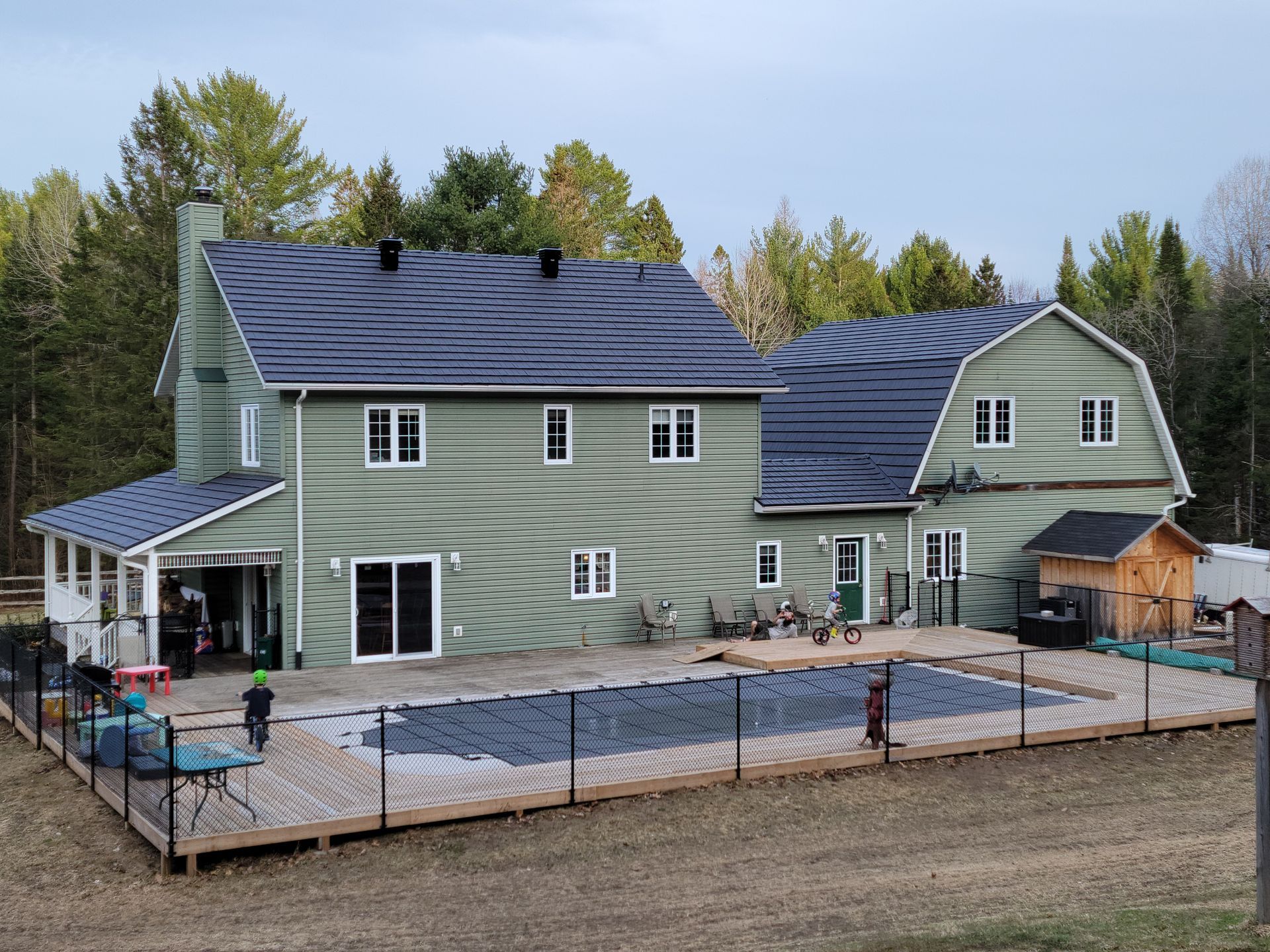 Green house with pool, black fence, and kids playing on wooden deck in wooded area.