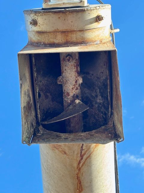 Close-up of a rusty, white grain auger's discharge spout with a rotating blade against a blue sky.