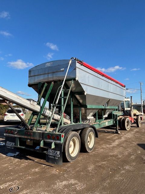 Large silver hopper trailer with red cover, green frame, and white wheels, parked outdoors on a sunny day.