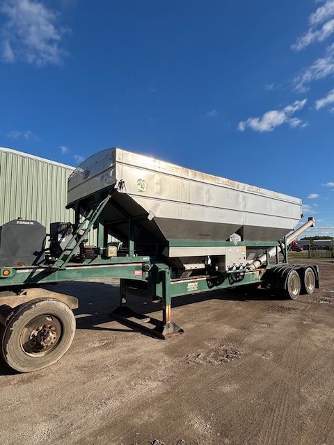 Silver and green agricultural fertilizer spreader trailer against a blue sky.