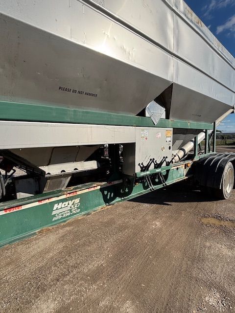 Green and silver agricultural fertilizer spreader trailer on a gravel road.