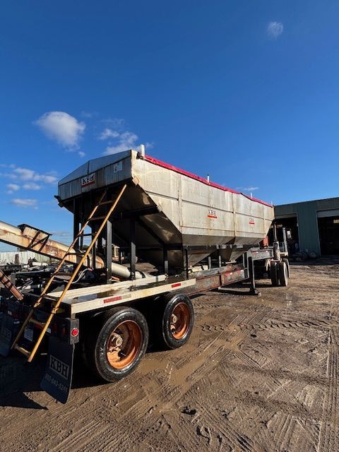 A large, silver hopper trailer with orange wheels sits outdoors under a blue sky.