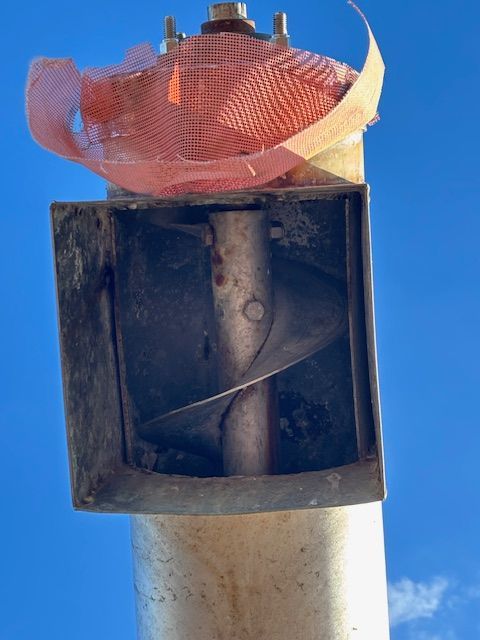 A metal auger with a spiral blade inside a square housing, with orange netting on top.