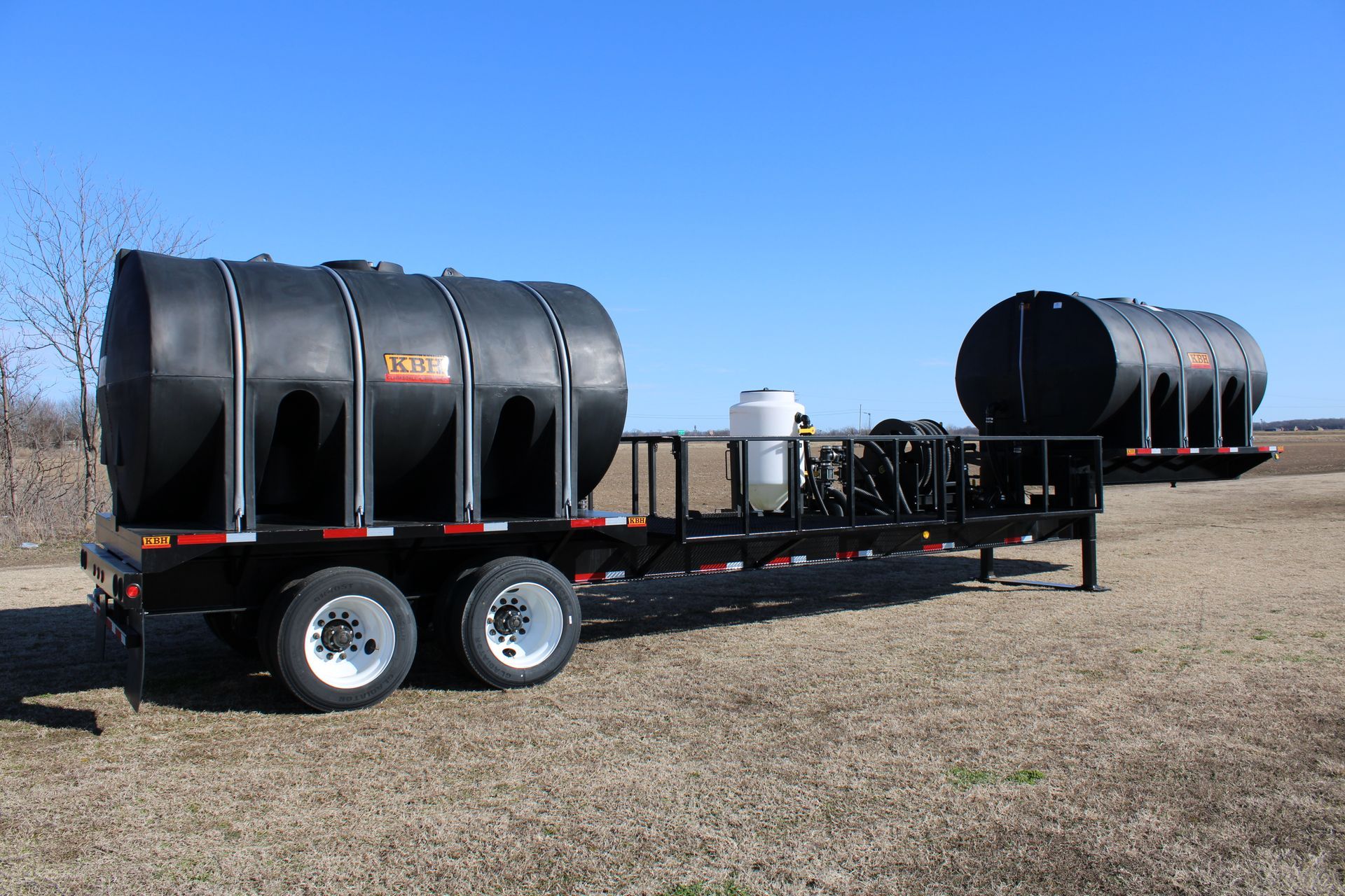 A black trailer carrying two large, black cylindrical tanks, parked outdoors on a sunny day.