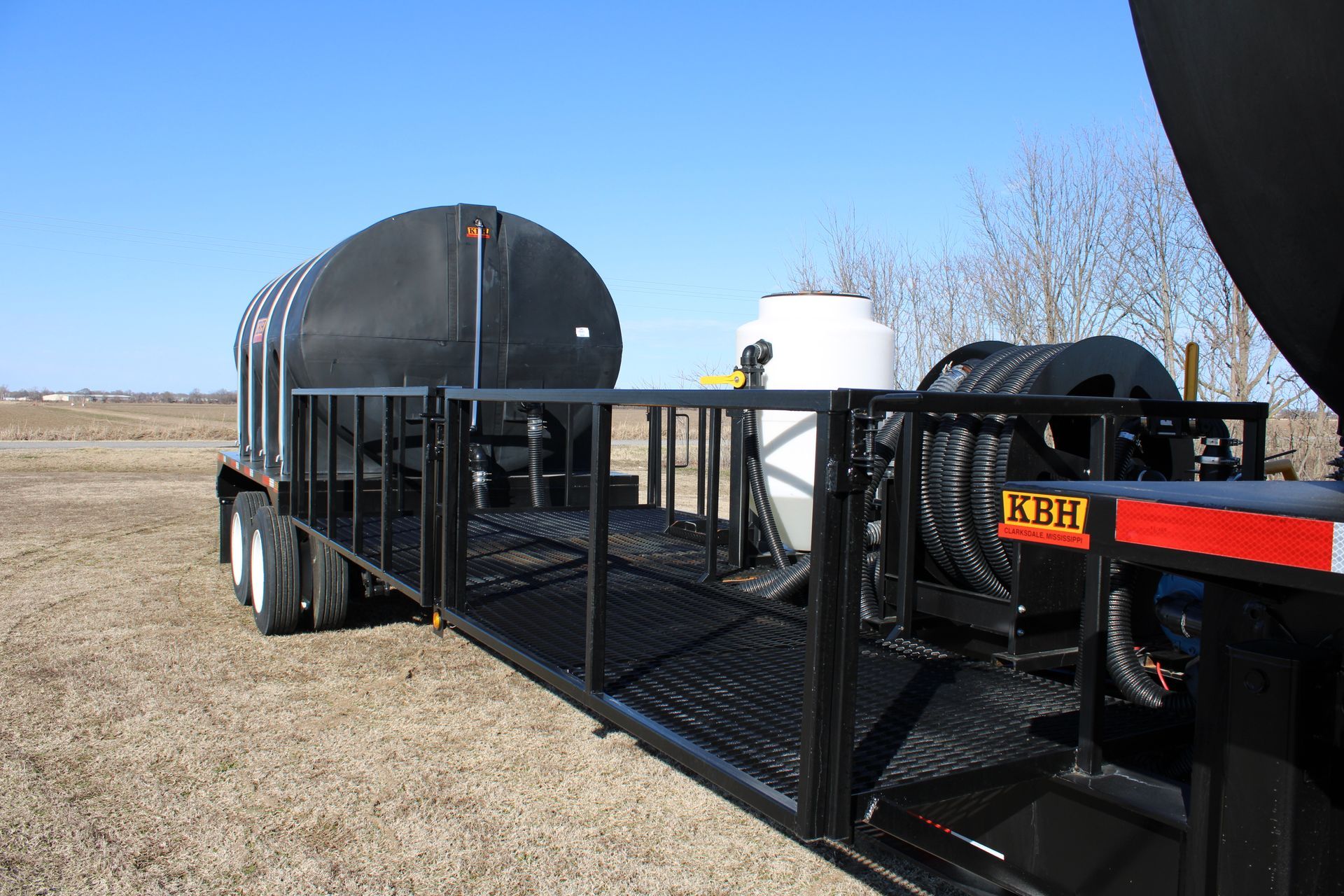 Black trailer with tanks and a hose reel in a field under a blue sky.