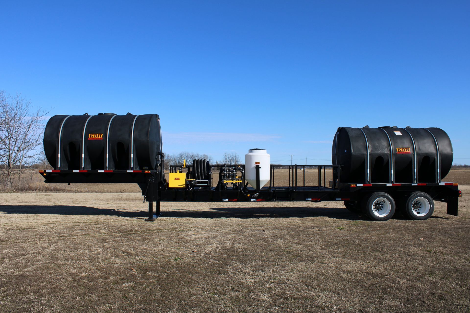 Black tanker trailer with two tanks and a pump system outdoors under a blue sky.