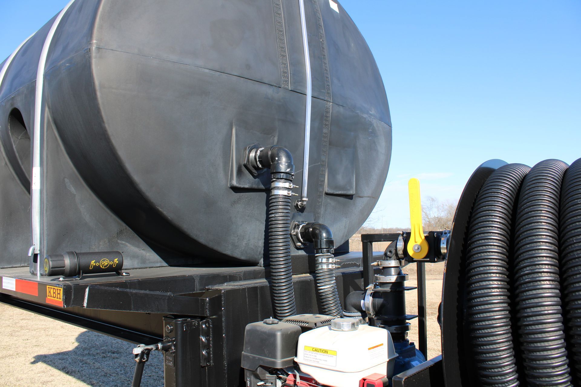 Black tank on a trailer with hoses and a gasoline engine, outdoors on a sunny day.
