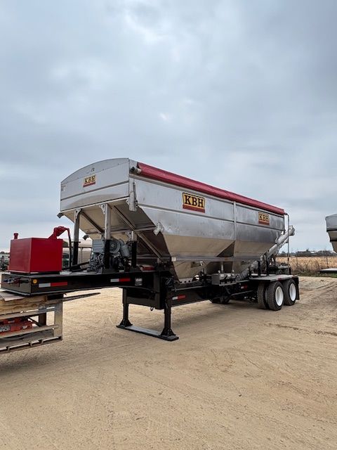 Silver grain trailer with red accents parked on gravel. Cloudy sky.