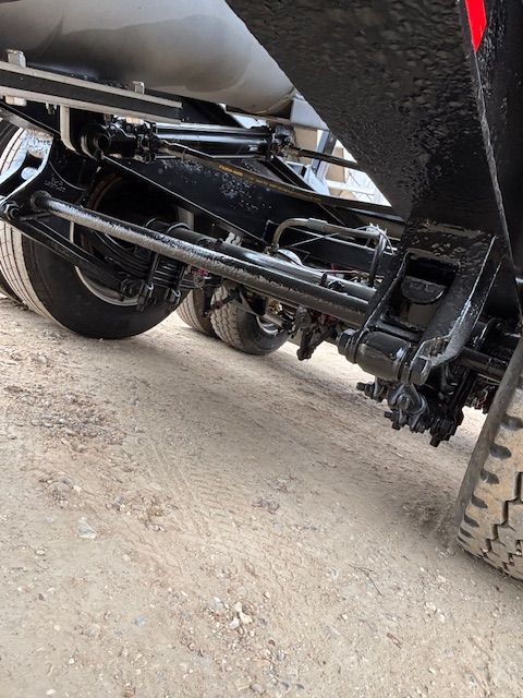 Underside view of a black truck chassis with wheels, suspension, and metal components on a dirt surface.