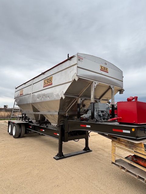 Silver and black agricultural grain trailer on a tan surface against a cloudy sky.