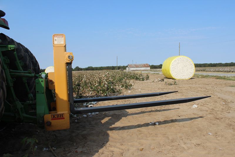 A tractor with a forklift attached to it is in a field with a bale of hay in the background.