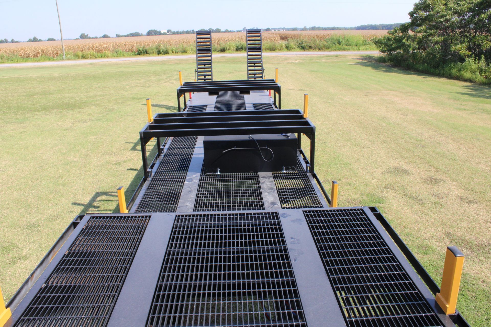 Metal trailer with ramps, grates, and yellow supports on a grassy field, with a cornfield in the background.