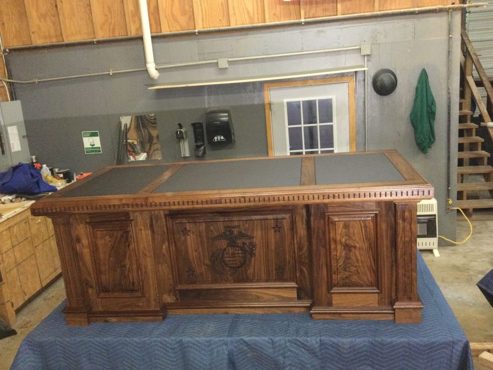 Wooden desk with a dark top, intricate carvings, and a crest. Set in a workshop with gray walls and a doorway.