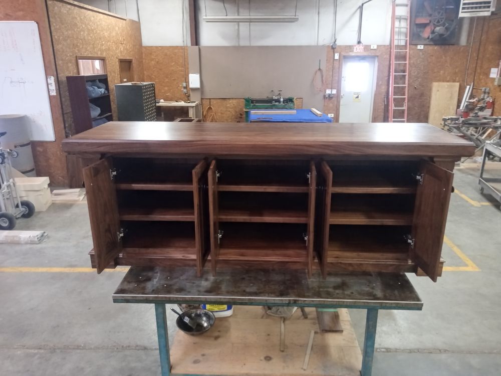 Wooden cabinet with three open doors, revealing shelves inside. It sits on a workbench in a workshop.