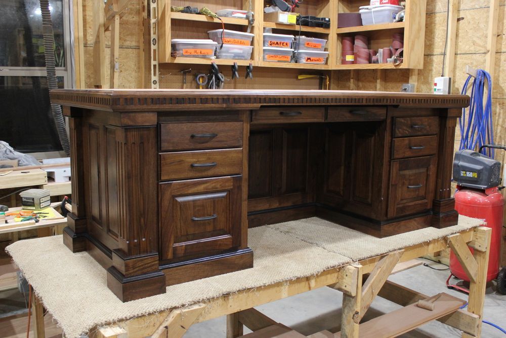 Wooden desk in workshop. Dark brown finish. Drawers on left and right, center space. Shelves and tools in background.