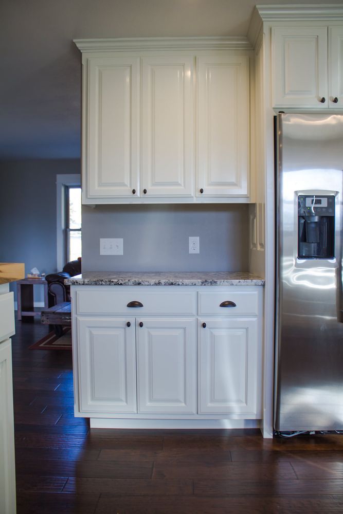 White kitchen cabinets with black hardware against a grey wall. Stainless steel refrigerator is to the right.