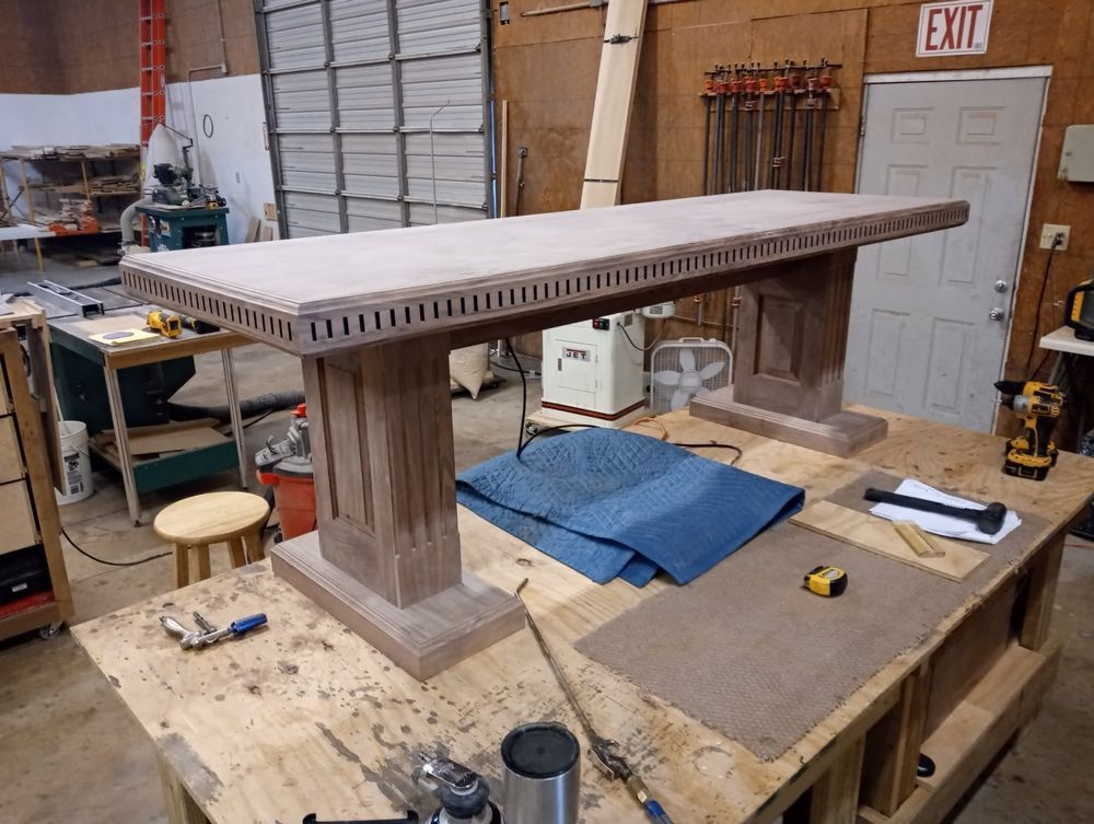 A partially built wooden table in a workshop, with a detailed carved edge and pedestal base.