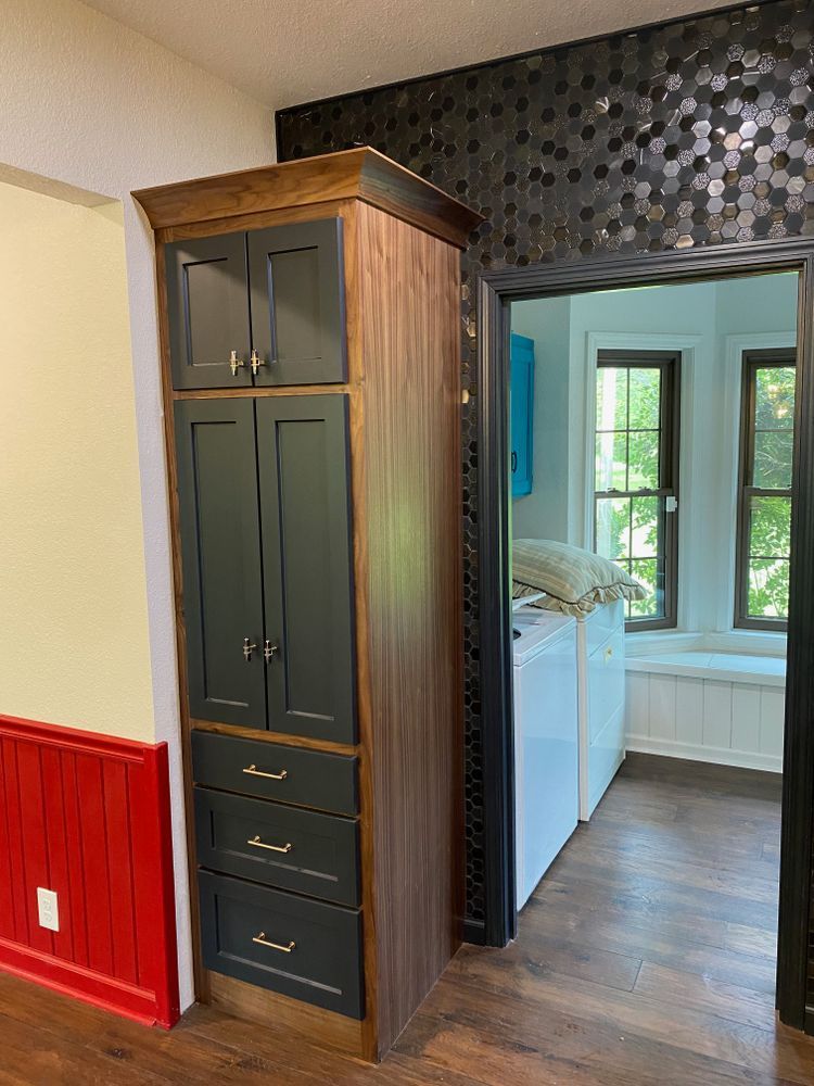 Tall wooden pantry cabinet with dark green doors and drawers, next to a black tiled wall and doorway.