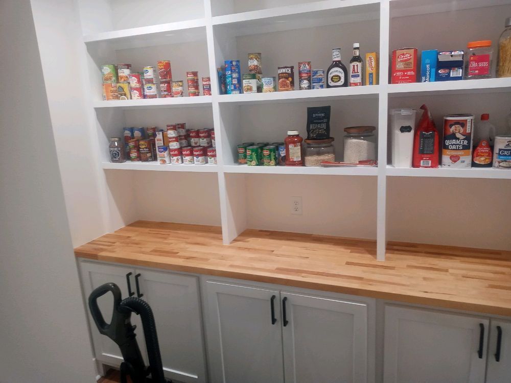 Pantry with white shelves filled with groceries, wood countertop, white cabinets below. A vacuum is leaning.