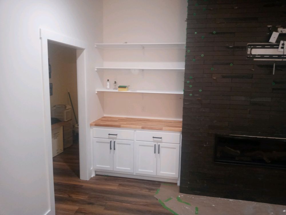 White built-in cabinet with butcher block counter and shelves; dark wall with fireplace to the right, doorway to the left.