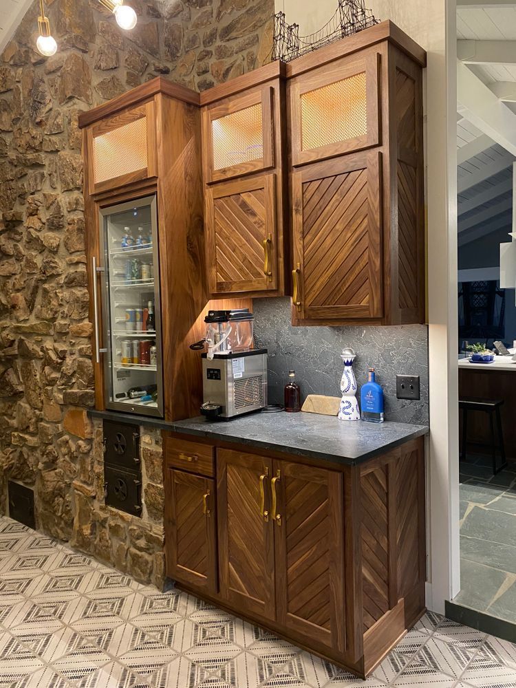 Built-in wooden bar with refrigerator, cabinets, and countertop against a stone wall and patterned floor.