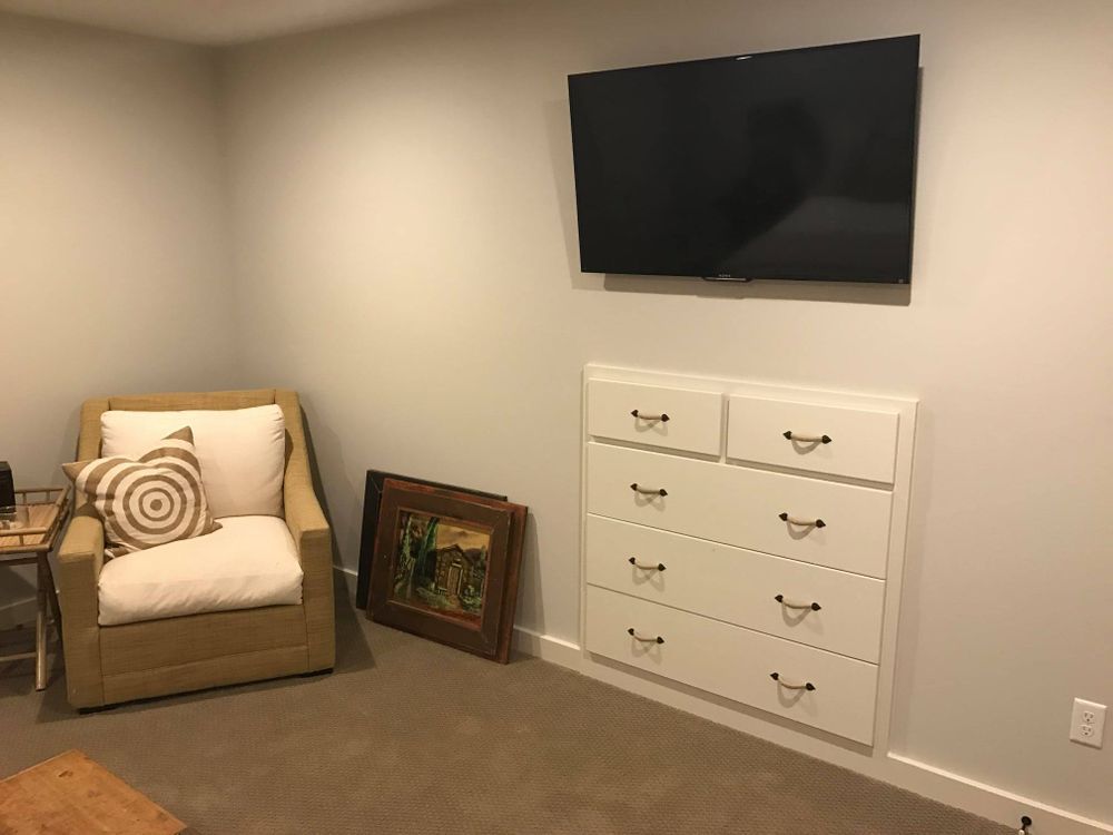 A beige armchair, white dresser, and TV on a gray wall in a room with a brown carpet.