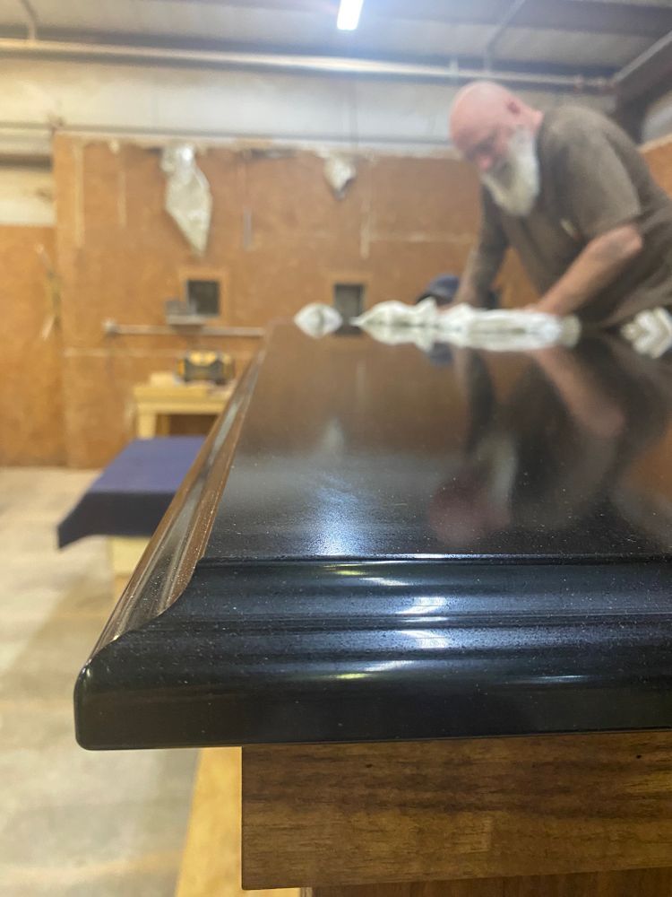 Man polishing dark, glossy table surface in a workshop. Focus on the table's edge.