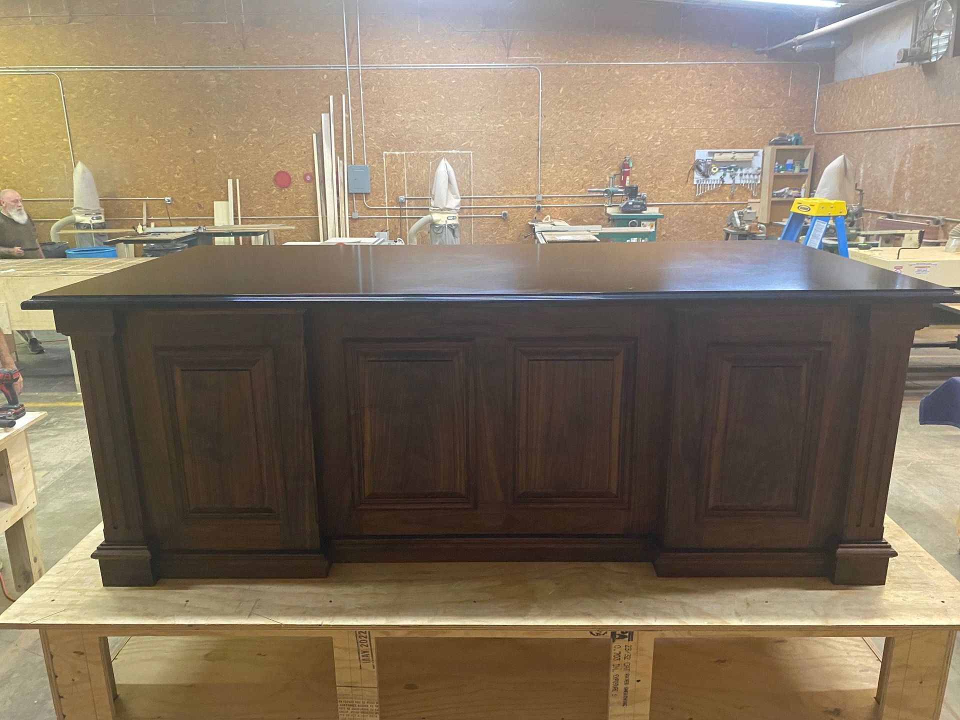 A large, dark wooden desk with paneling sits on a wooden work table in a workshop.