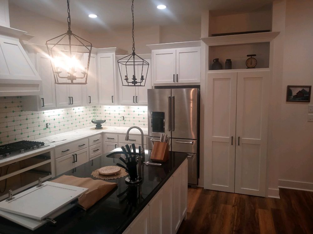 White kitchen with island and stainless steel refrigerator. Black countertops and dark metal light fixtures.