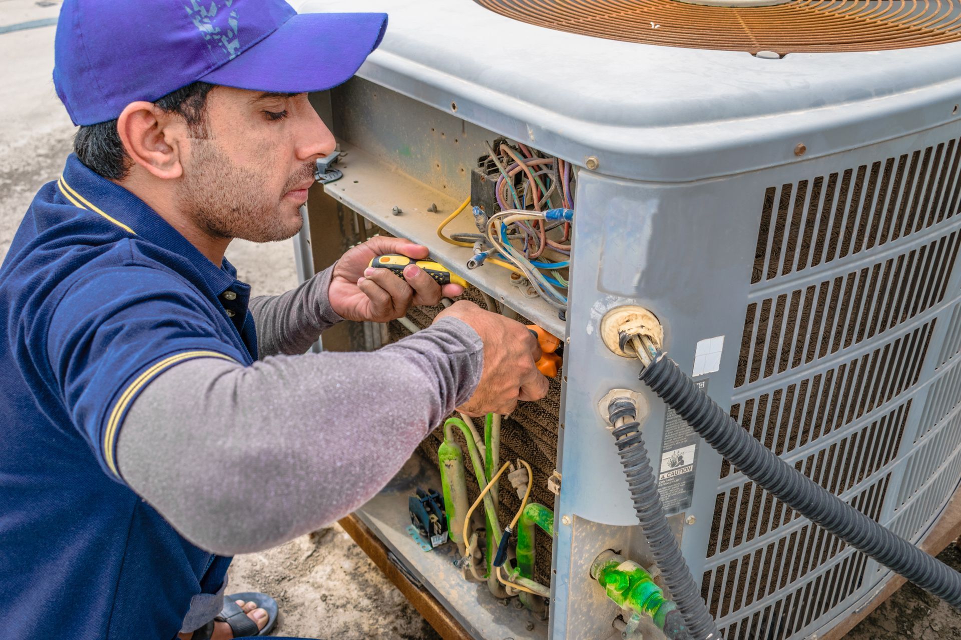 Technician in a purple cap works on exposed wiring inside an outdoor HVAC unit.