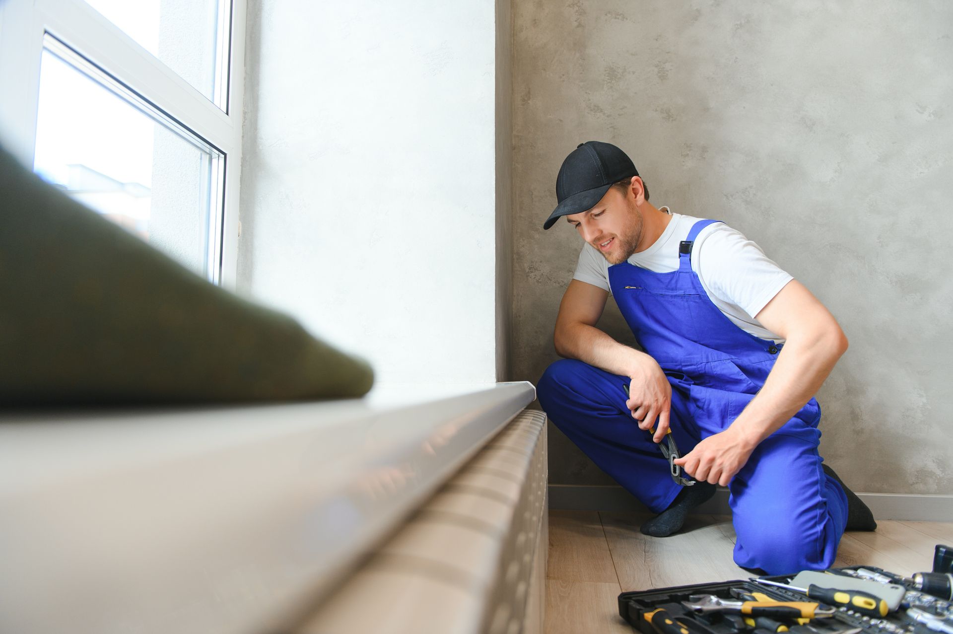 Worker in blue overalls inspecting a wall near a window with tools on the floor.