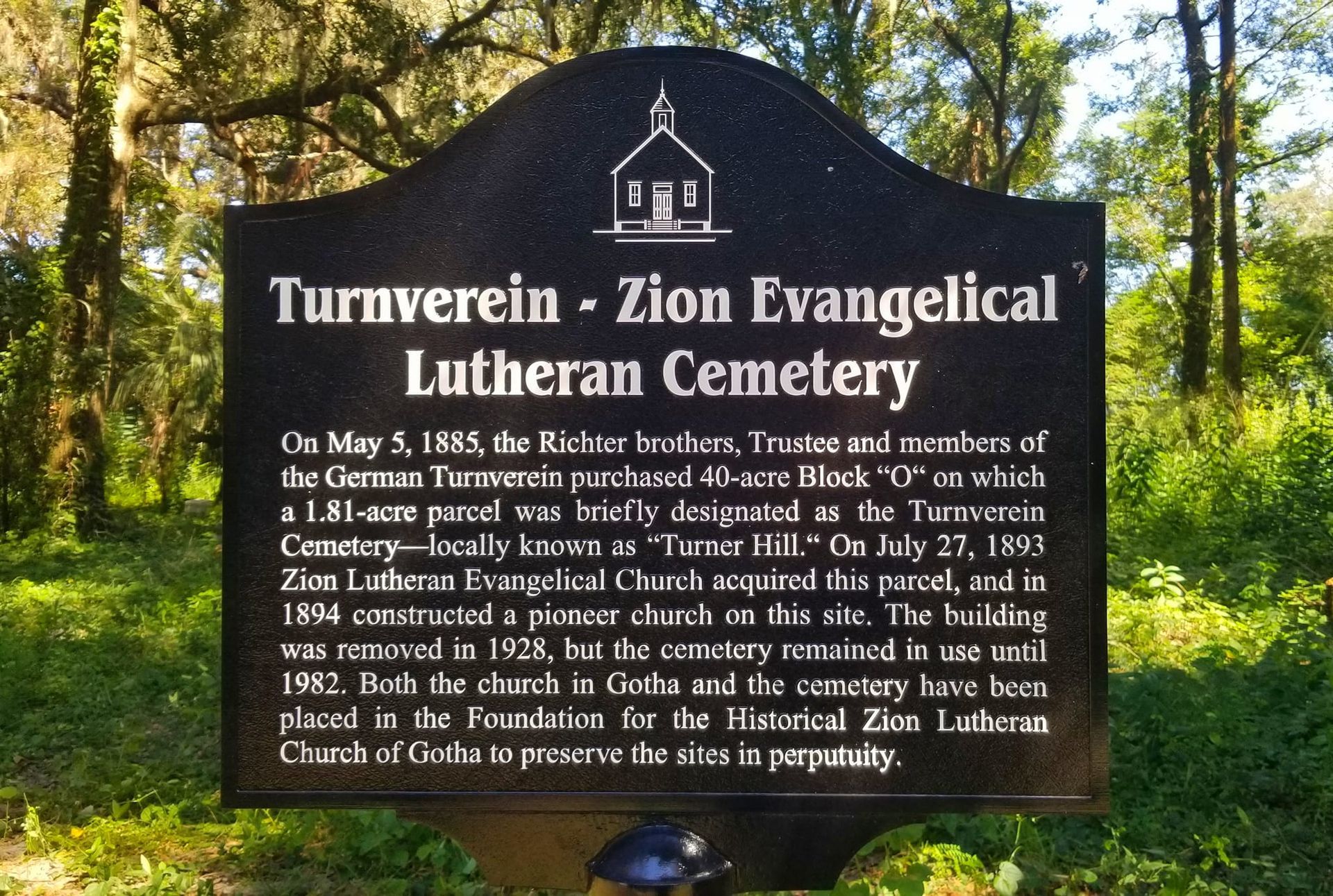 Sign for Turnverein-Zion Evangelical Lutheran Cemetery; black with white text and church graphic, in a natural setting.