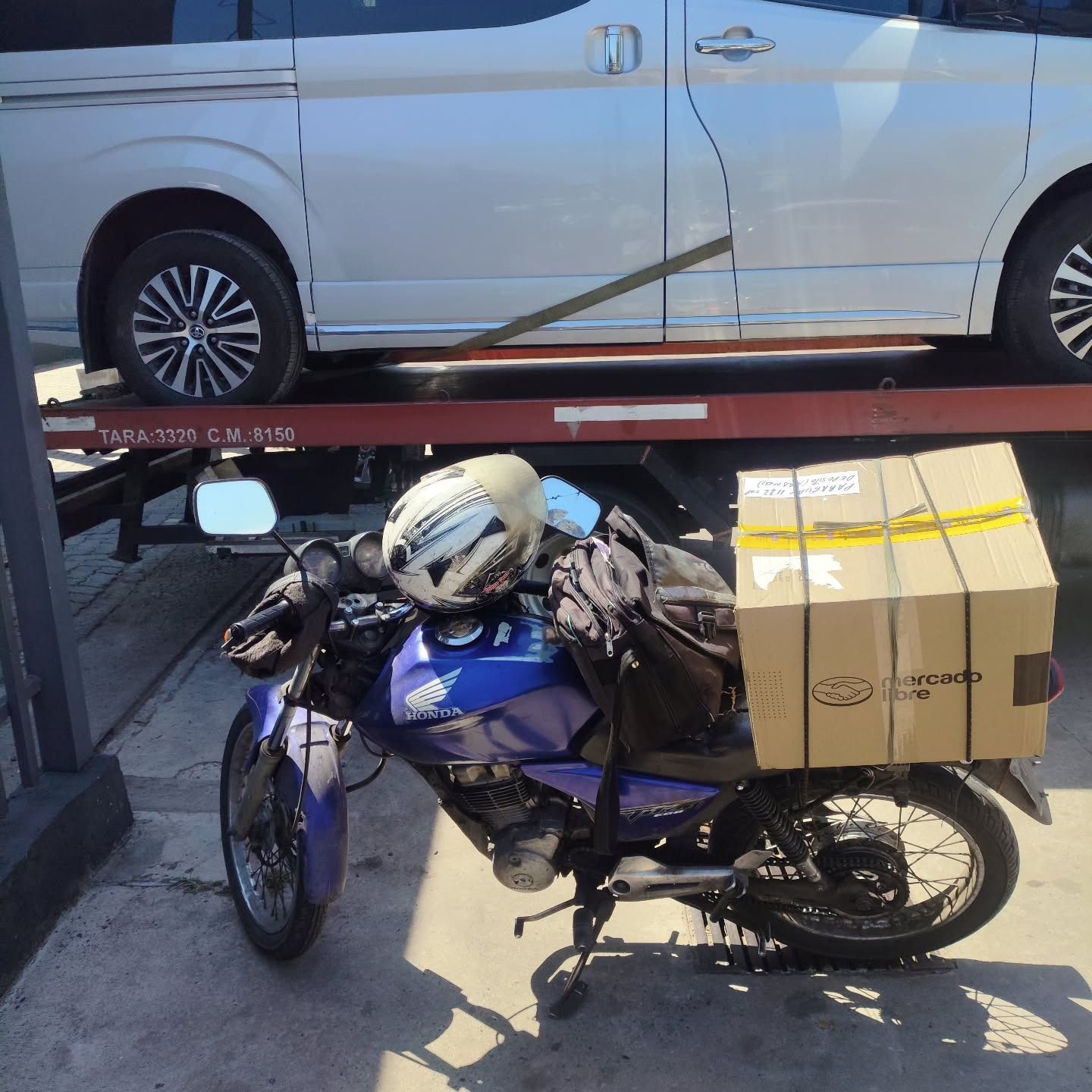 Una motocicleta cargada de cajas y bolsas estaba estacionada junto a una furgoneta blanca bajo la luz del sol.