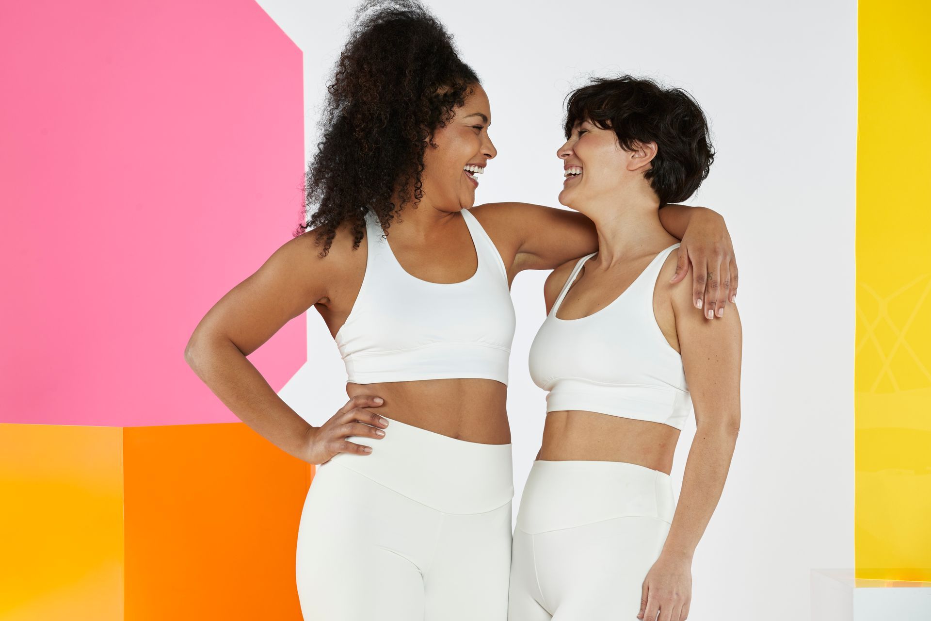 Two women in white sports bras and shorts are standing next to each other.